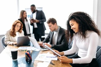 Happy attractive female employee is sitting in office at business meeting, learning financial graphs. Beautiful young adult business woman doing paperwork, smiling